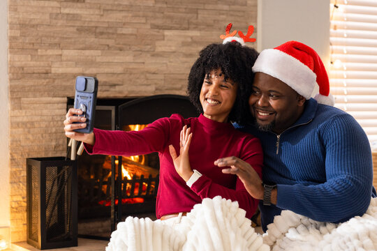 Diverse couple taking selfie on sofa in front of fireplace holding smartphone under string lights