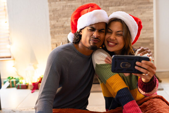 Couple taking selfie leaning on plush rug in living room with smartphone, Santa hats and fireplace