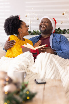 Diverse couple laughing on sofa under blanket in Christmas living room holding red hardback book