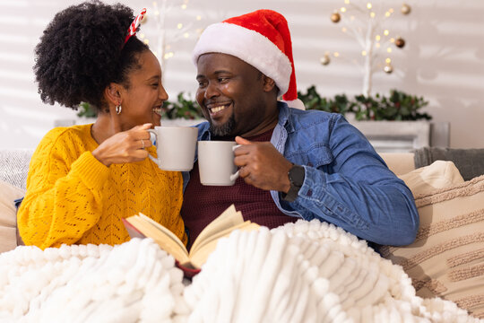African American couple sipping from matching mugs on couch wearing Santa hat with string lights