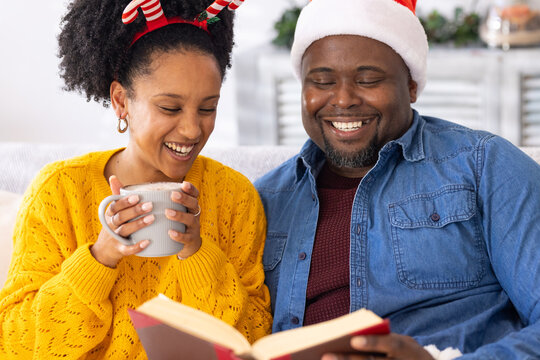 Diverse couple wearing Santa hat, antler headband while reading red hardcover book on sofa with mug