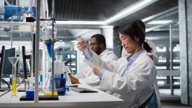 Assay development specialist uses pipette to transfer liquid into test tube containing blue solution at laboratory bench. Asian lab engineer does pipetting procedure with chemical reagent, camera A