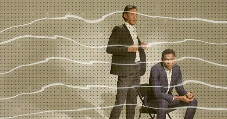 Posing male models in suits adjusting jacket lapels, clasping hands on folding chair in studio