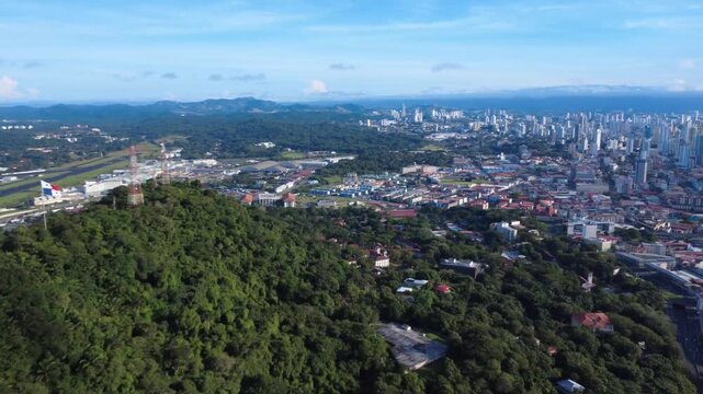 Panama flag flies high over lush green hill overlooking city . Aerial Shot of ancon hill, with Panama City in the back. 