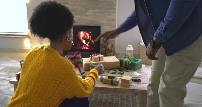 With man entering wearing Santa hat, diverse couple wrapping holiday gifts on floor by fireplace