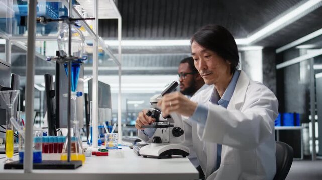 Clinical microbiologist examining microscope glass slide in laboratory to view cells. Asian lab worker checks sample slide before imaging process using optical device, camera A