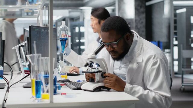 Clinical microbiologist operating compound microscope does detailed sample observation, recording findings. Biotechnology expert studies tissue under optical device, documenting results, camera B