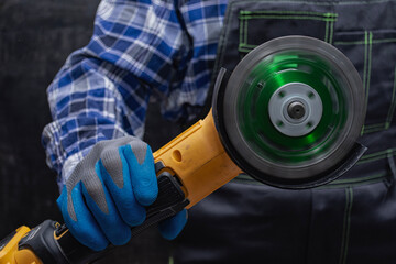 Construction worker in a blue checkered shirt and gloves operates a yellow angle grinder with a rotating concrete cutting disc. Power tool for construction repairs.