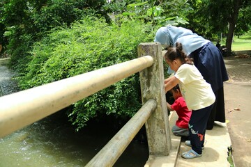 A woman and two children lean over a railing to view a river in a lush park. Bright day, casual clothing, and masks highlight family bonding and outdoor exploration.