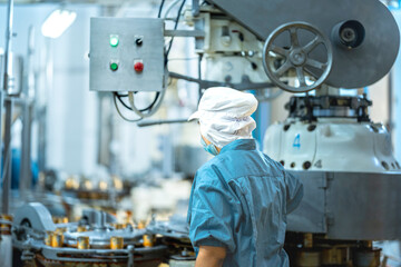 A factory worker in a blue uniform, mask, and cap monitors an automated production line. Canned goods are moving on a conveyor belt in a food processing plant.