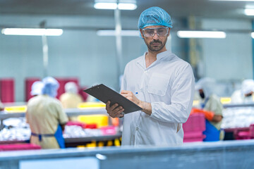 A male factory inspector in a lab coat, hairnet, and safety glasses stands and observes a busy food...