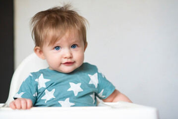 Smiling baby boy in a blue T-shirt sitting on a white high chair in a white kitchen
