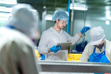 A male factory inspector in a lab coat, hairnet, and safety glasses is checking inventory. He holds...