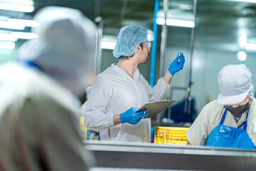 A male inspector in a lab coat, hairnet, and safety glasses checks a small fish. He is holding a clipboard and doing a quality control check in a food processing factory.