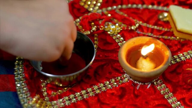 A hand performing a Hindu ritual with a lit diya and red powder on a decorative red cloth.
