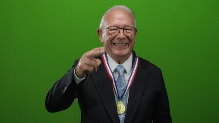 Senior businessman wearing a suit smiles confidently against an isolated green background, showcasing a victory medal around his neck with a proud salute.