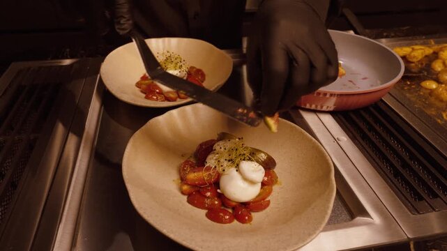 A chef wearing black gloves carefully arranges a gourmet dish with burrata and tomatoes in a modern kitchen, showcasing fine dining plating art.