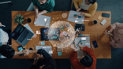 A vibrant team collaborates around a table filled with laptops, notebooks, and data charts, fostering innovation and strategic discussions in a modern office setting. Trope