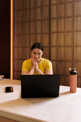 Person in a yellow shirt smiles while using a laptop in a cozy indoor workspace during the afternoon