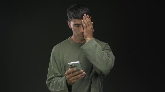 Man in olive green sweater holding silver smartphone in studio with dark green background and covering face with hand while frowning; worry.