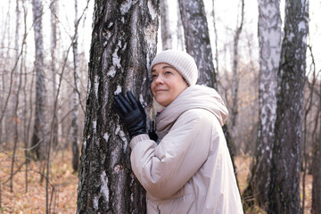 Middle-aged woman hugs tree in park. Spiritual healing, love of nature. Energy recharge in the forest. Happy adult woman hugs tree and smiles during autumn season outdoors. Natural lifes