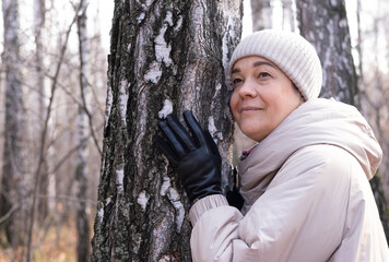 Middle-aged woman hugs tree in park. Spiritual healing, love of nature. Energy recharge in the forest. Happy adult woman hugs tree and smiles during autumn season outdoors. Natural lifes
