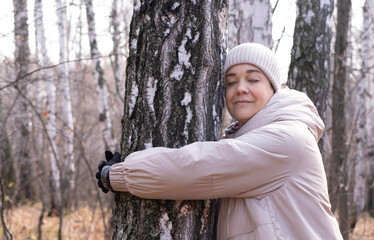 Middle-aged woman hugs tree in park. Spiritual healing, love of nature. Energy recharge in the forest. Happy adult woman hugs tree and smiles during autumn season outdoors. Natural lifes