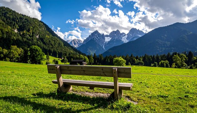A wooden bench sits in a grassy field with views of tall mountains under a partly cloudy sky. The sun casts shadows