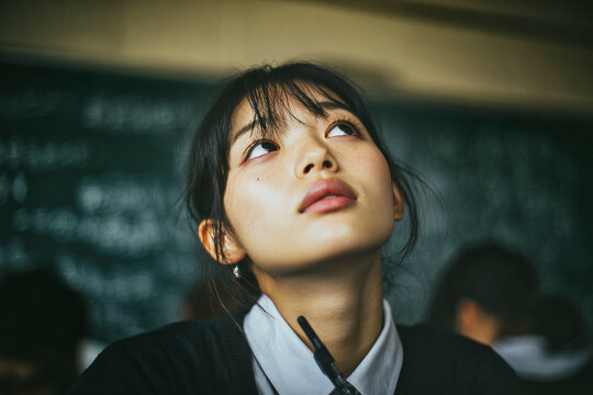 Portrait of a thoughtful schoolgirl holding a pen and looking upward in front of a blackboard in a classroom with natural light