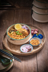 Beautiful still life of traditional Northern Thai Khao Soi chicken curry noodles served in an enamel bowl on a wooden table, with crispy noodles, shallots, lime, chili oil.