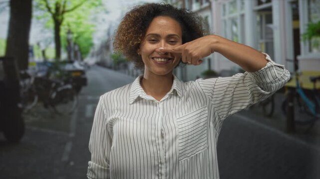 Woman points finger to nose while smiling on a city street with parked bicycles and row houses; playful confidence.