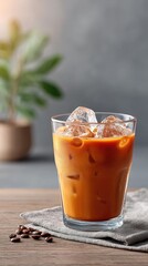Refreshing iced coffee in a clear glass with ice cubes and coffee beans on a textured surface with a blurred green plant in the background and warm sunlight