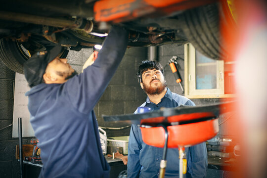 Two mechanics are focused on inspecting a vehicle that is elevated, with one shining a light on the underside. The workshop setting includes visible tools and equipment.
