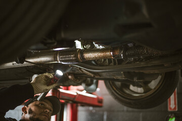 A mechanic holds a flashlight while inspecting the underside of a car in a garage. The environment suggests a typical automotive repair setting.