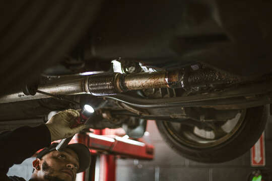 A mechanic holds a flashlight while inspecting the underside of a car in a garage. The environment suggests a typical automotive repair setting.