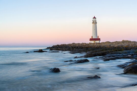 Lighthouse at sunset on rocky coast with ocean waves and pastel sky long exposure