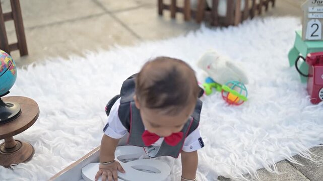 An adorable baby crawling on a soft white rug in an outdoor photoshoot setup, surrounded by decorative props such as a vintage telephone, wooden fence, stuffed toy, Eiffel Tower model, and date calend