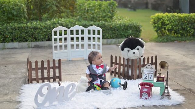 An adorable baby crawling on a soft white rug in an outdoor photoshoot setup, surrounded by decorative props such as a vintage telephone, wooden fence, stuffed toy, Eiffel Tower model, and date calend