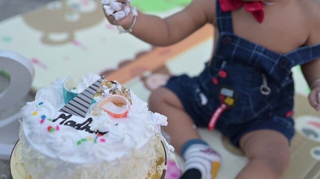 A cute baby celebrating a first birthday with a cake smash during an outdoor photoshoot. The child is sitting on a colorful play mat next to a decorated birthday cake and a &ldquo;One&rdquo; sign, wearing a cute 
