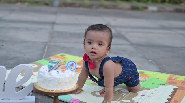 A cute baby celebrating a first birthday with a cake smash during an outdoor photoshoot. The child is sitting on a colorful play mat next to a decorated birthday cake and a &ldquo;One&rdquo; sign, wearing a cute 