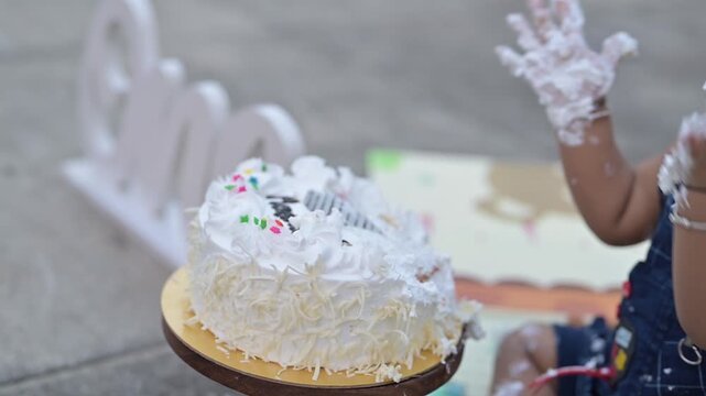 A cute baby celebrating a first birthday with a cake smash during an outdoor photoshoot. The child is sitting on a colorful play mat next to a decorated birthday cake and a &ldquo;One&rdquo; sign, wearing a cute 