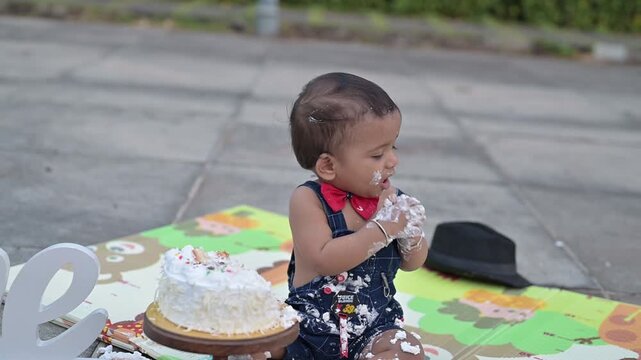 A cute baby celebrating a first birthday with a cake smash during an outdoor photoshoot. The child is sitting on a colorful play mat next to a decorated birthday cake and a &ldquo;One&rdquo; sign, wearing a cute 