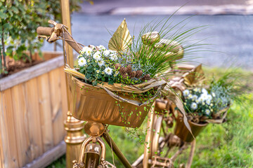 Ein zauberhaftes, ganz in Gold getauchtes Fahrrad steht als strahlendes Symbol der ewigen Liebe auf sattgrünem Rasen. Jeder Korb ist ein kleines Nest aus frischem Grün, zarten weißen Blüten und liebev