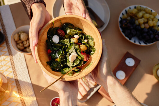 Unrecognizable woman handing wooden bowl of fresh salad with spinach, tomatoes, mozzarella and chickpeas to man during outdoor meal, hands of both visible above table with food - Powered by Adobe