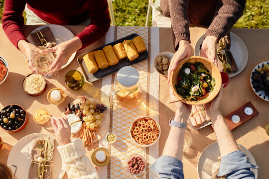 From above view of family sharing food and drinks at outdoor table, passing salad bowl, eating corn, enjoying meal together, hands visible