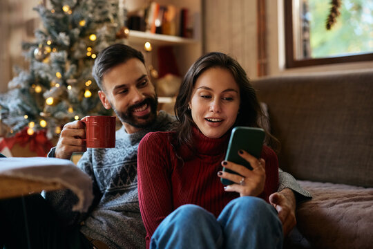 Young couple using smart phone while relaxing at home on Christmas day. - Powered by Adobe