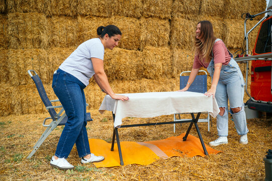Two women setting a table near orange camper and hay bales.