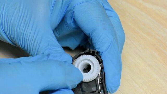 Blue-gloved hands perform precise gear maintenance inside a printer cartridge-clean handling, smooth rotation, and focused technical care highlight the repair process