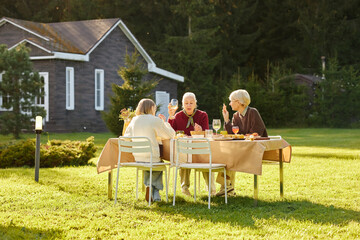 Senior and middle-aged women and girl sitting outdoors at table enjoying conversation and drinks in...