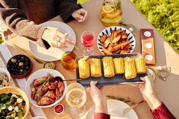 Two women enjoying outdoor picnic, serving grilled corn and chicken wings, sharing food and drinks...